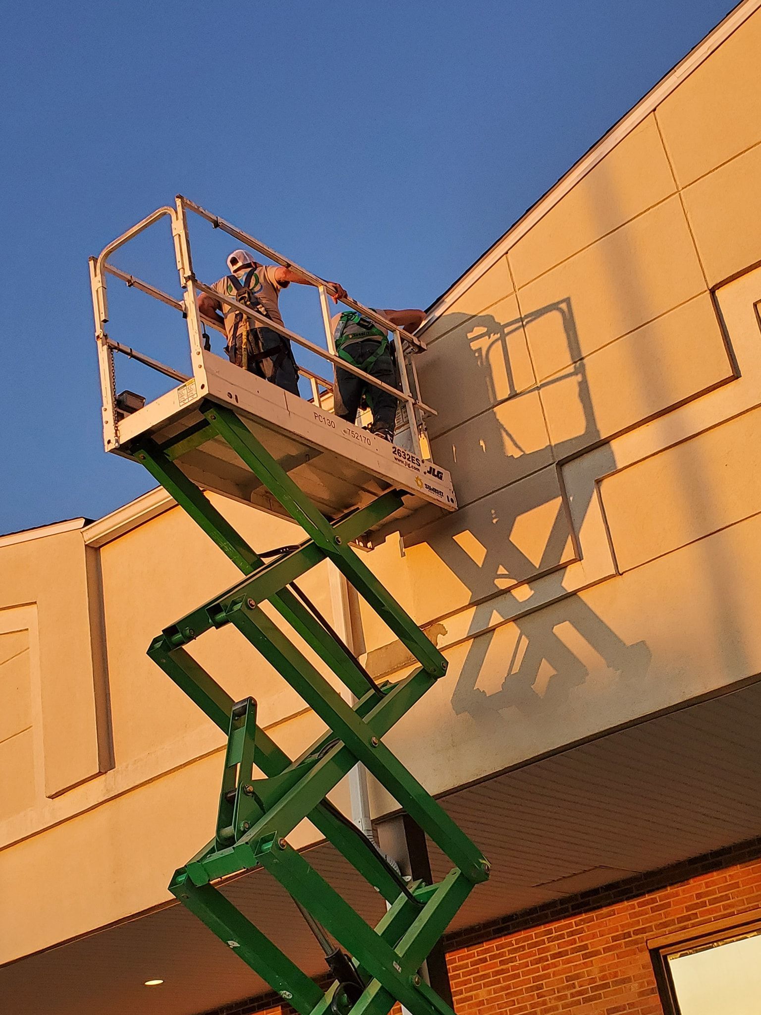 Two workers on a lift repair a building's exterior under a blue sky; the lift's shadow is cast on the wall.