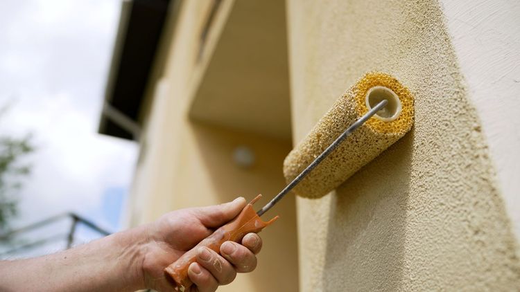 Hand painting a textured, beige exterior wall with a paint roller.