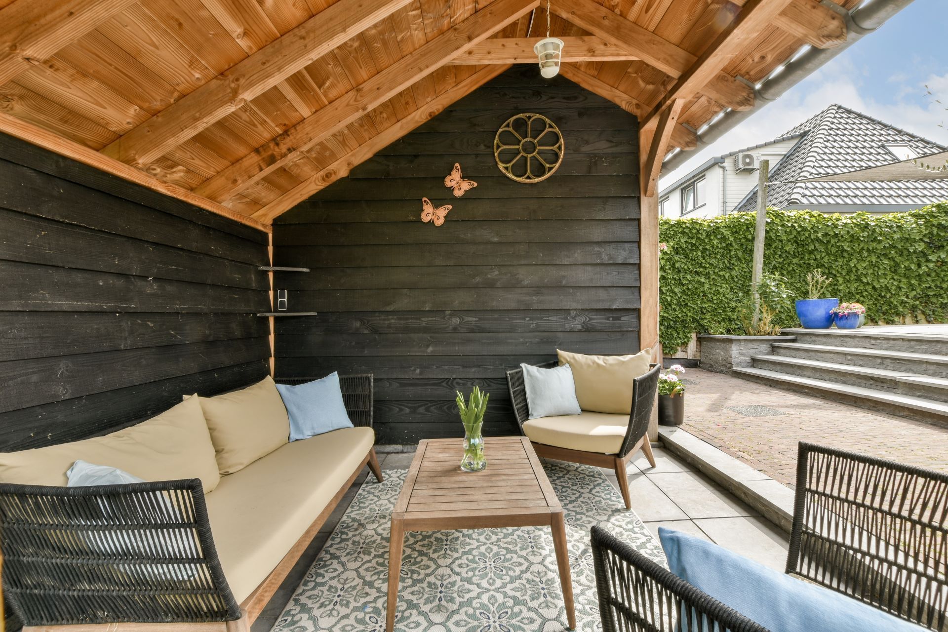 Outdoor covered seating area with a couch, chairs, and a wooden table; black walls, wooden ceiling, and a garden view.