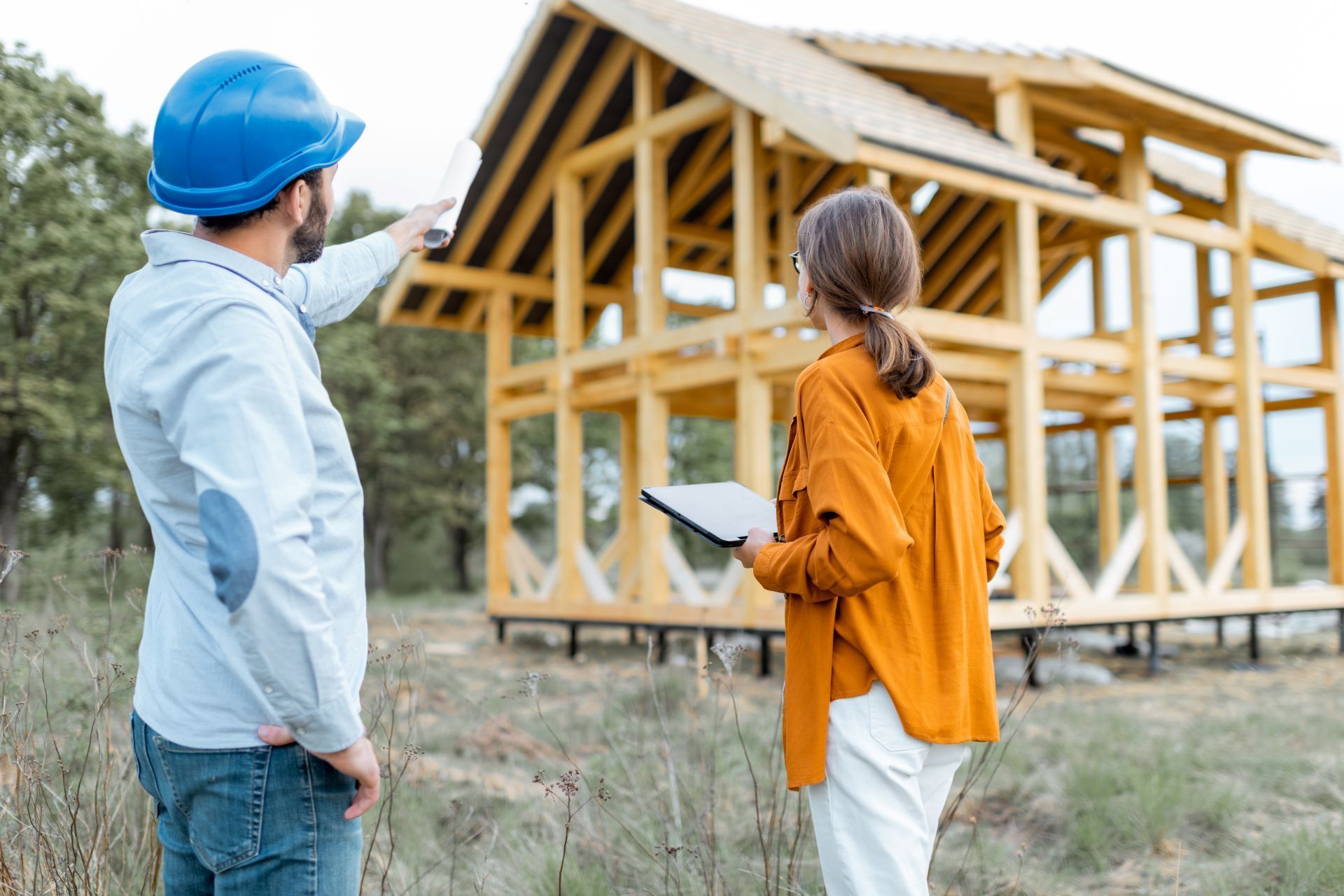 Construction workers review blueprints at a cabin's wooden frame. The man wears a blue helmet, pointing. The woman holds a tablet.