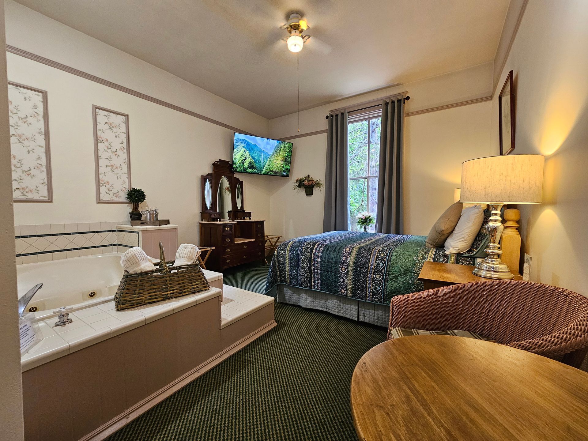 A hotel room with a bed , chair , table and jacuzzi tub.