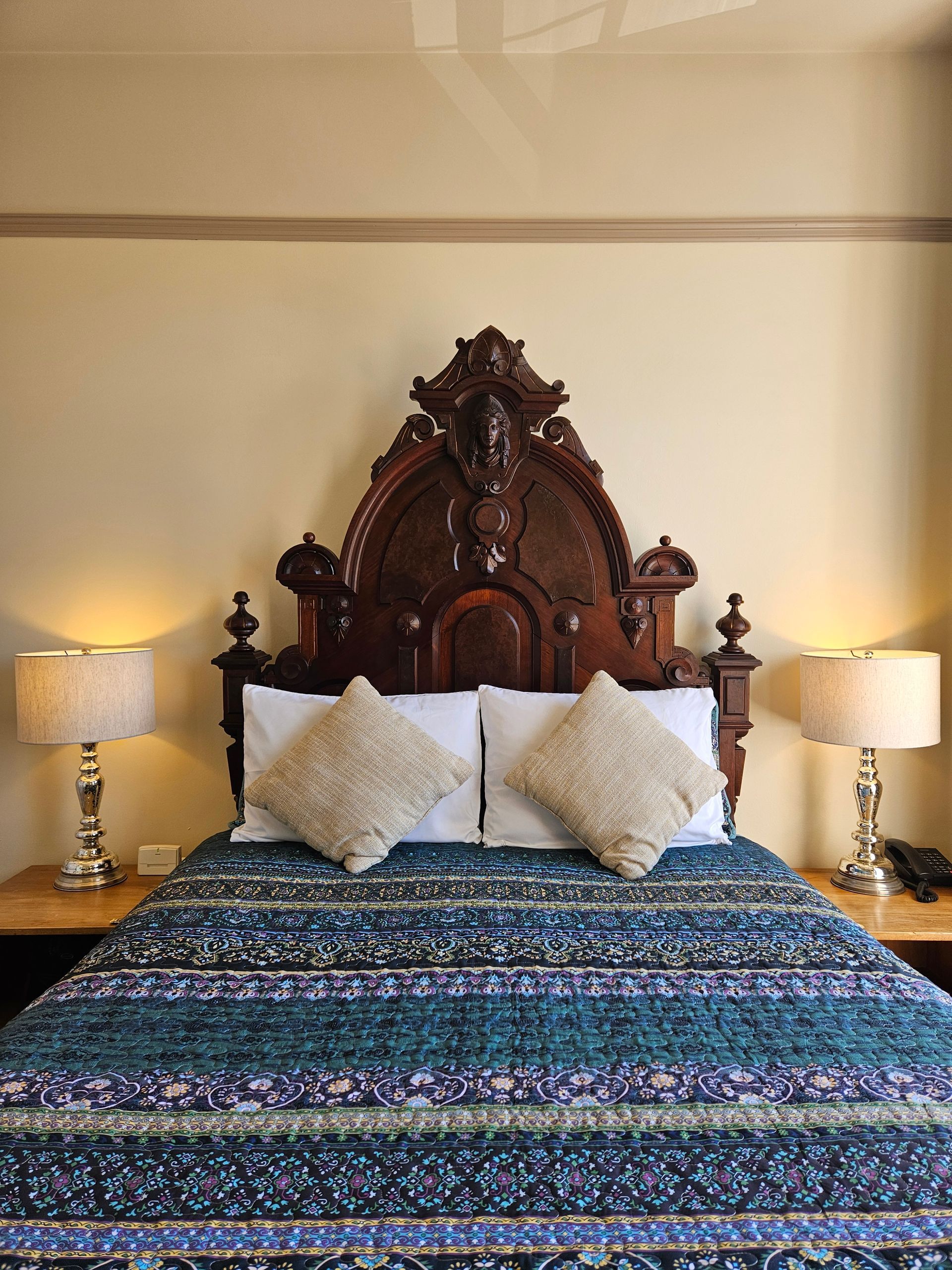 A large wood carved antique bed with a woman's face carved at the top and two side tables with lamps on them