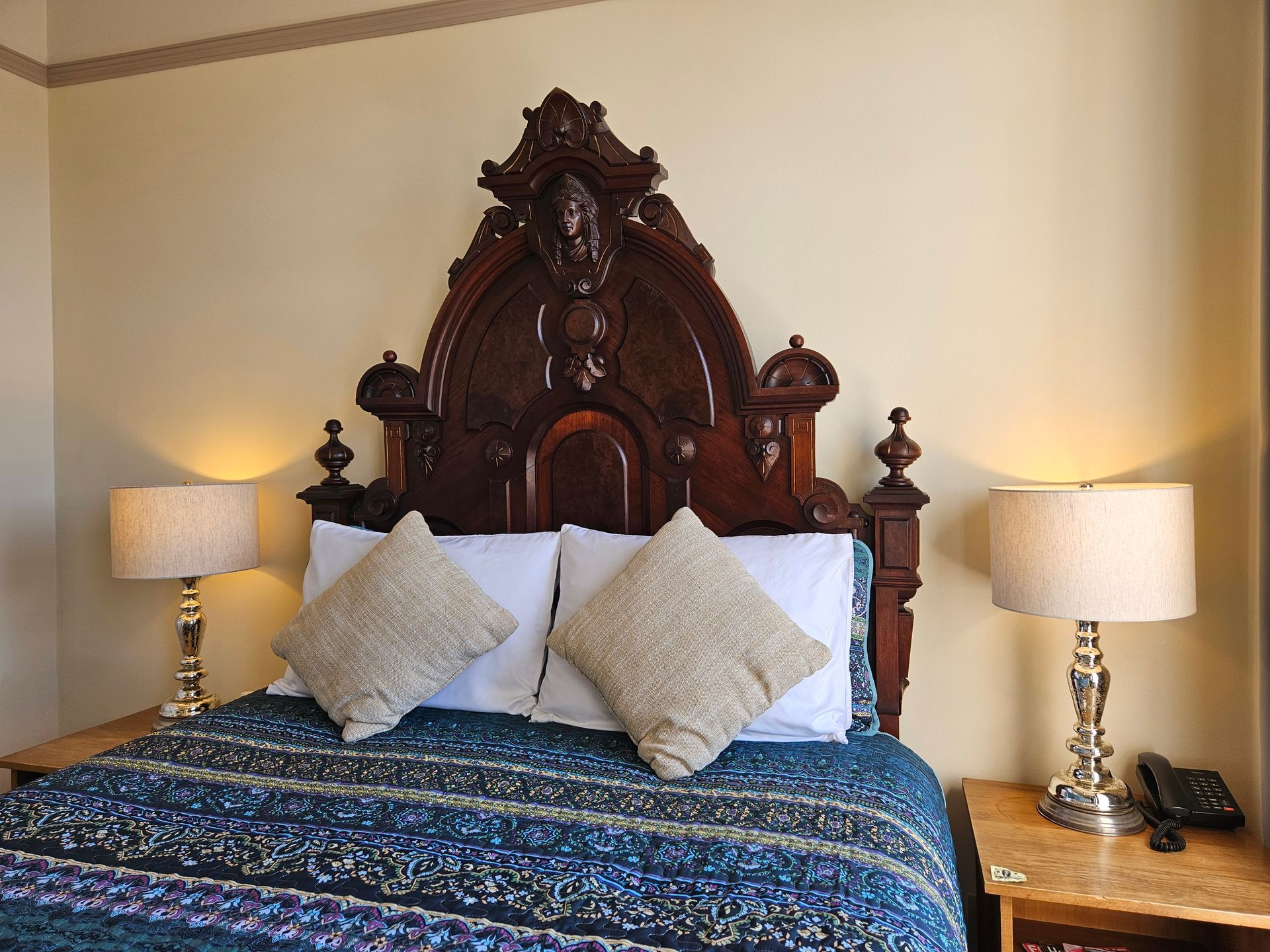 Head board of a antique wooden bed with a woman's face carved in it and two lamps on top of two side tables.