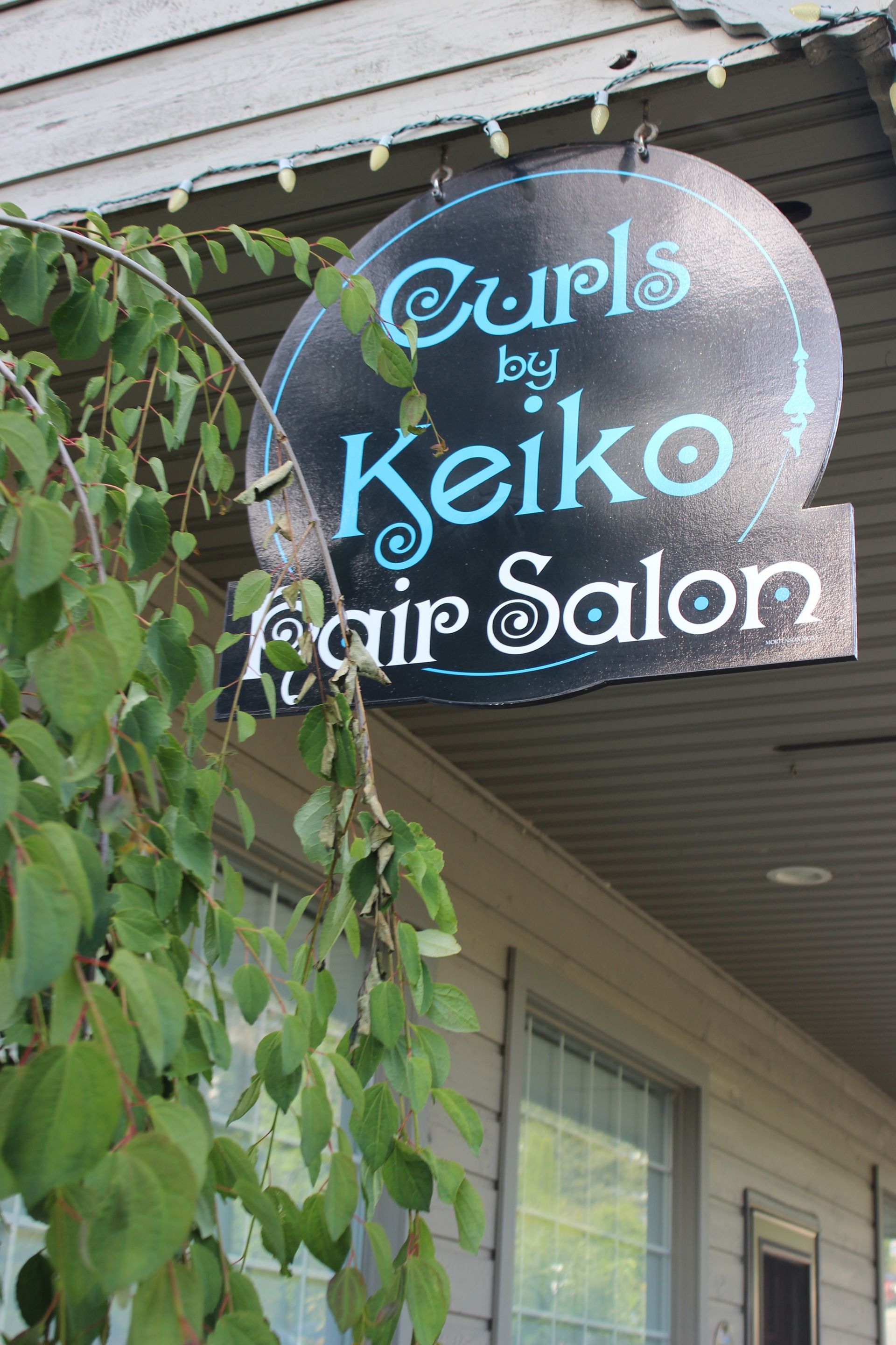 A sign for curls by keiko hair salon hangs from the side of a building.