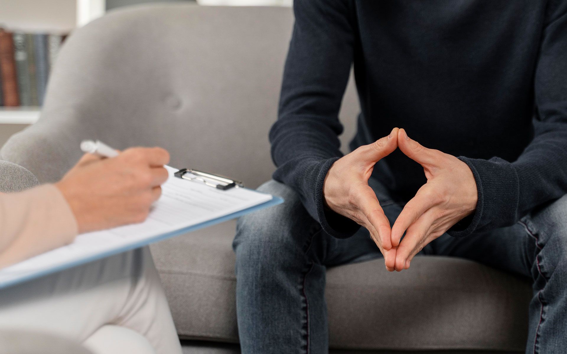 A man is sitting on a couch talking to a woman who is writing on a clipboard.