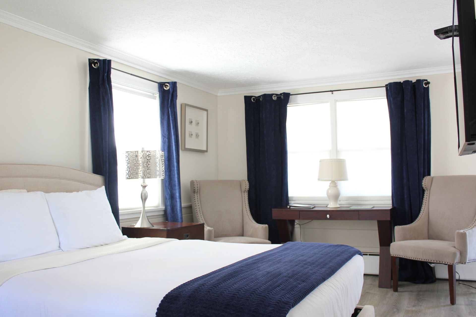 Bedroom with a white bed, navy blue accents, two windows with navy curtains, and two beige armchairs.