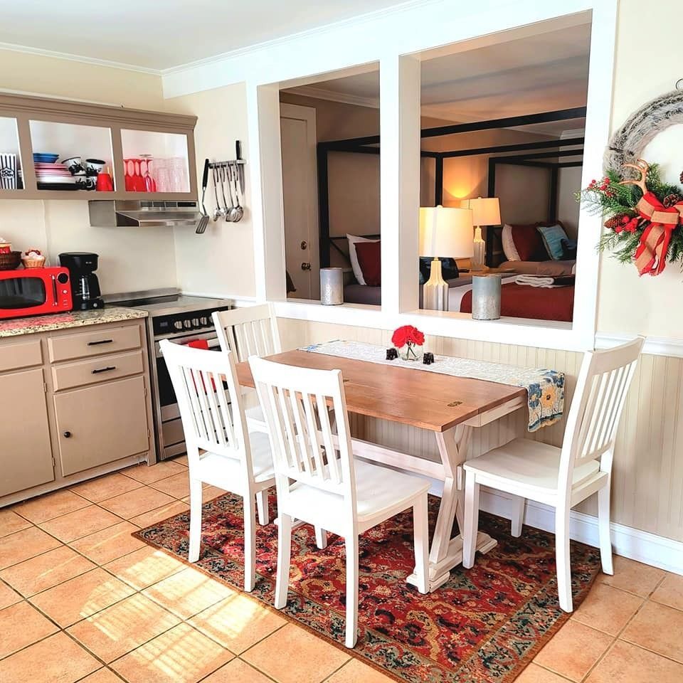 Kitchen with small table and chairs, visible through an opening to a bedroom; red, white and beige colors.