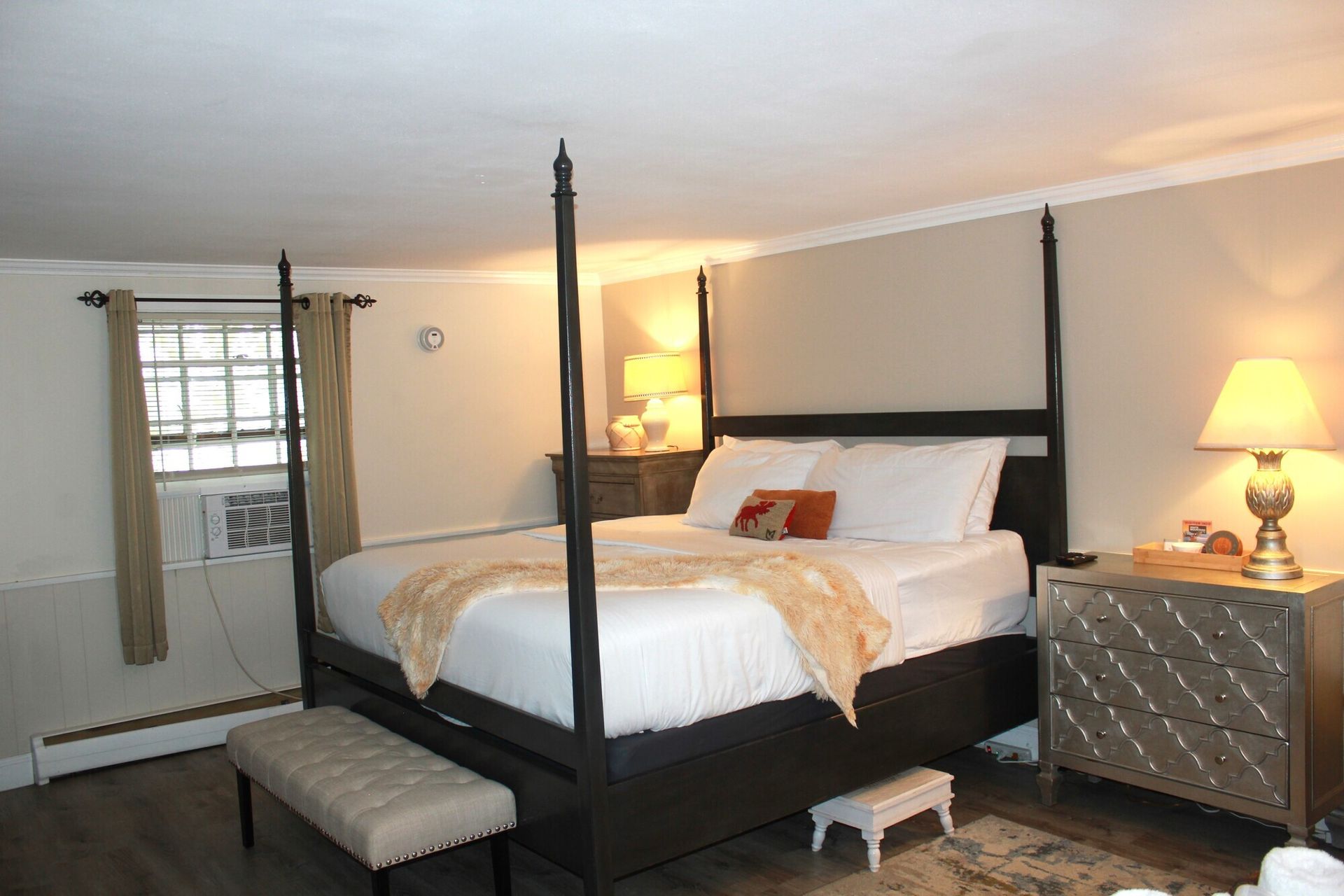 Bedroom with a dark four-poster bed, nightstands, a bench, and a window with an AC unit.