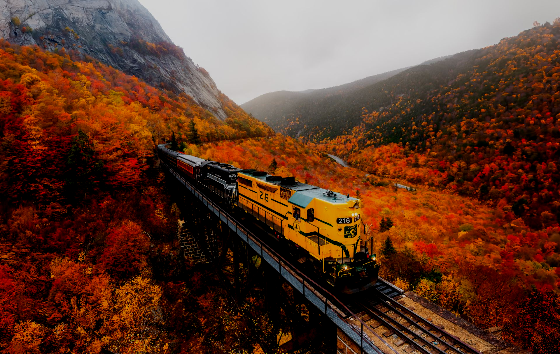 Yellow train travels through autumn foliage along a mountain rail.