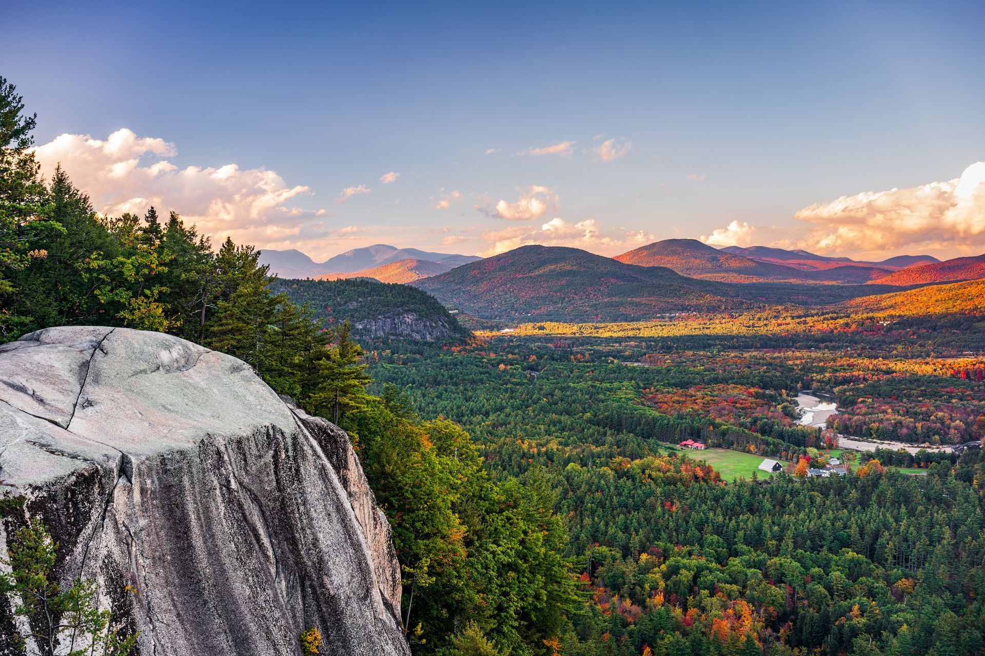 Rocky overlook of a forested valley and distant mountains under a colorful sky.