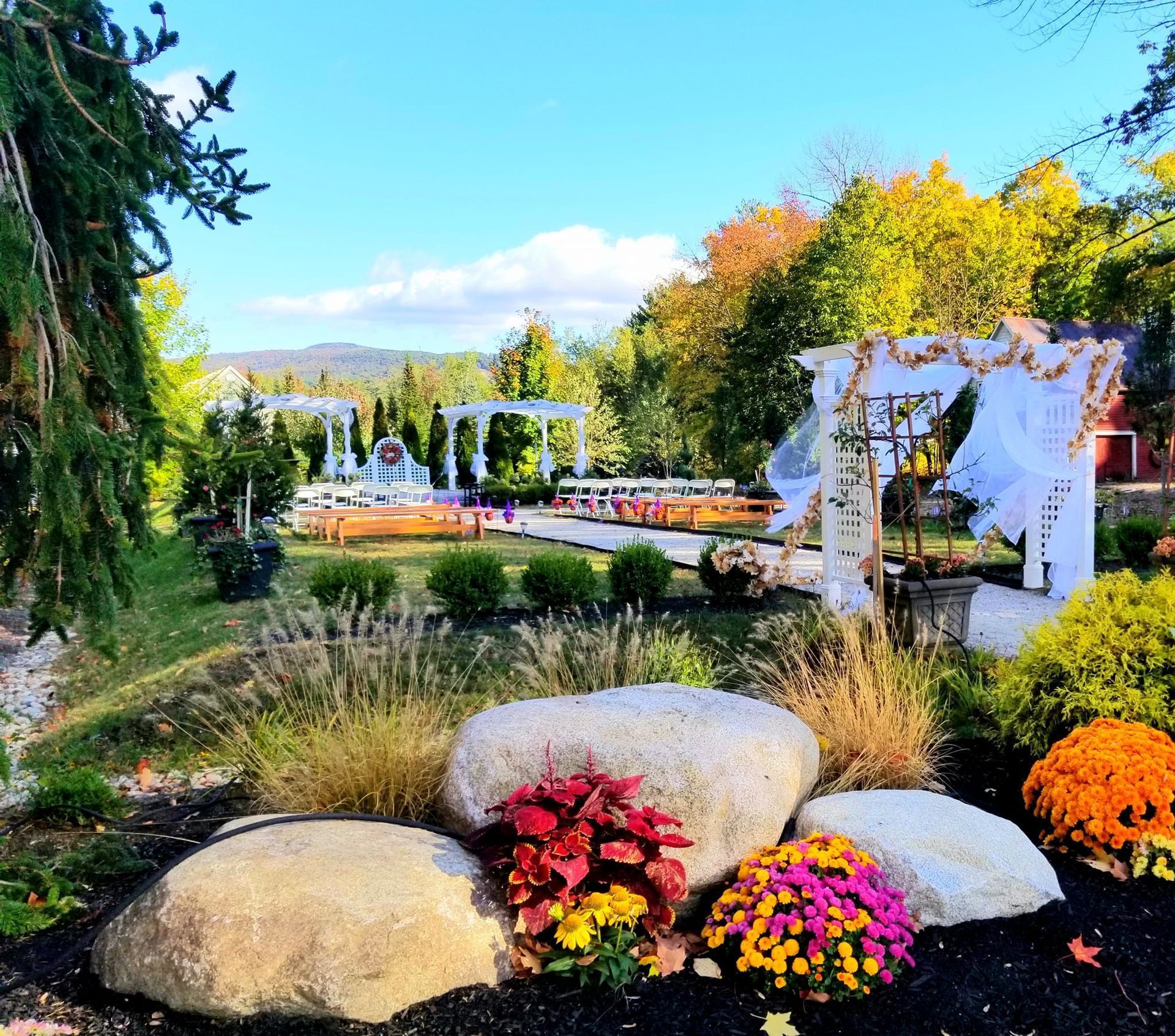 Wedding venue with white arches, tables, and decorations in a garden setting, with large rocks and autumn foliage.