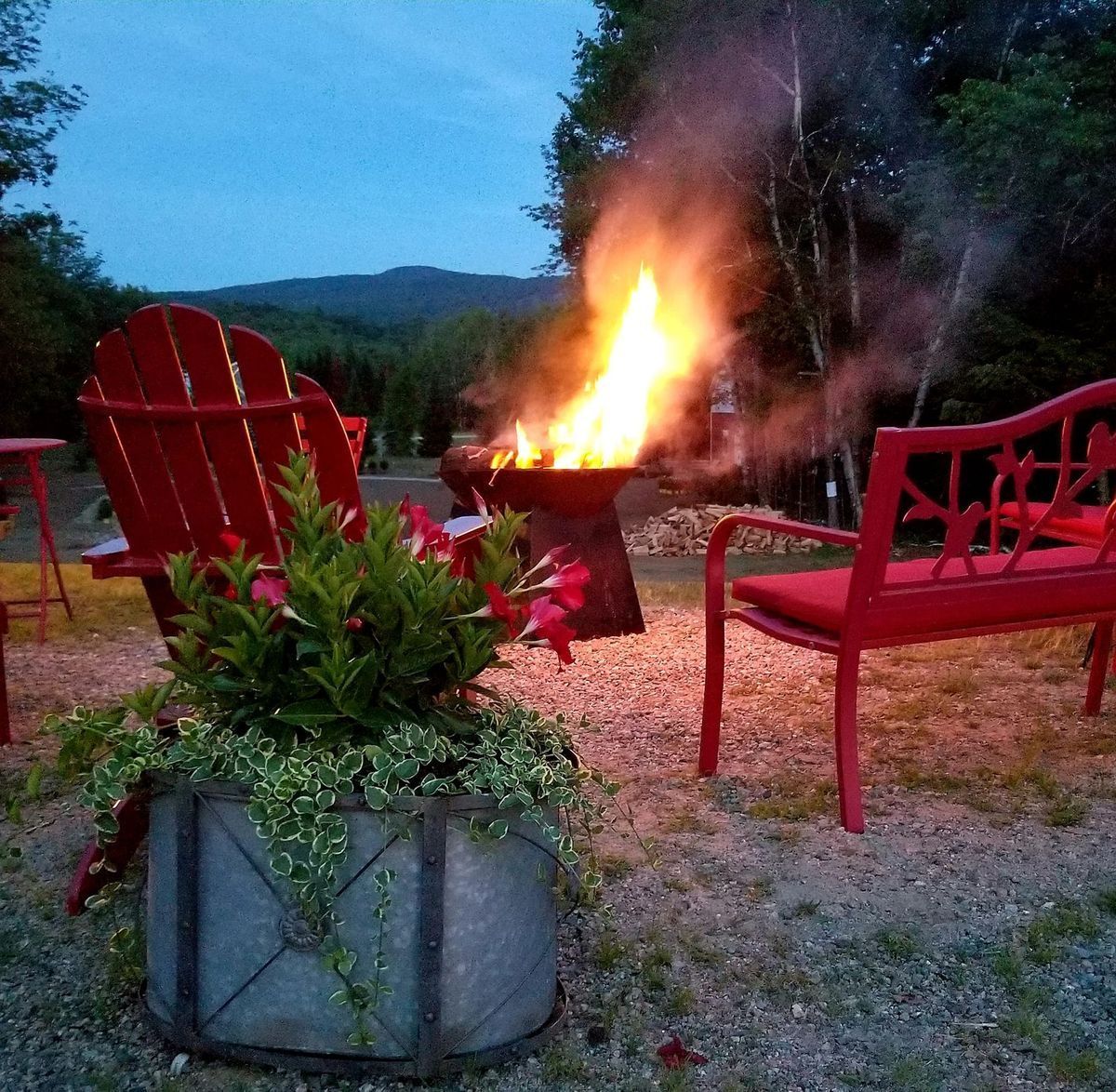 Red chairs surround a fire pit with a large fire, in an outdoor setting at dusk, with plants in a container.