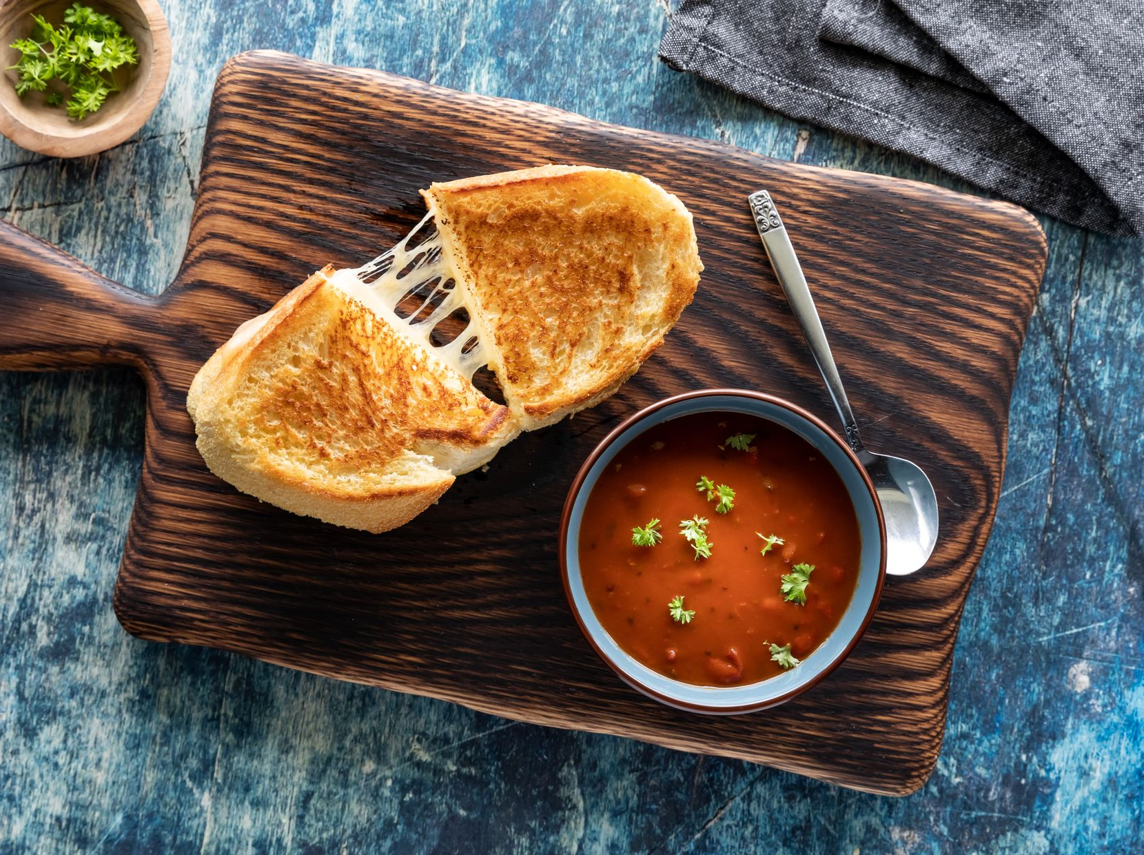 Grilled cheese sandwich with melted cheese next to a bowl of tomato soup, on a wooden board.