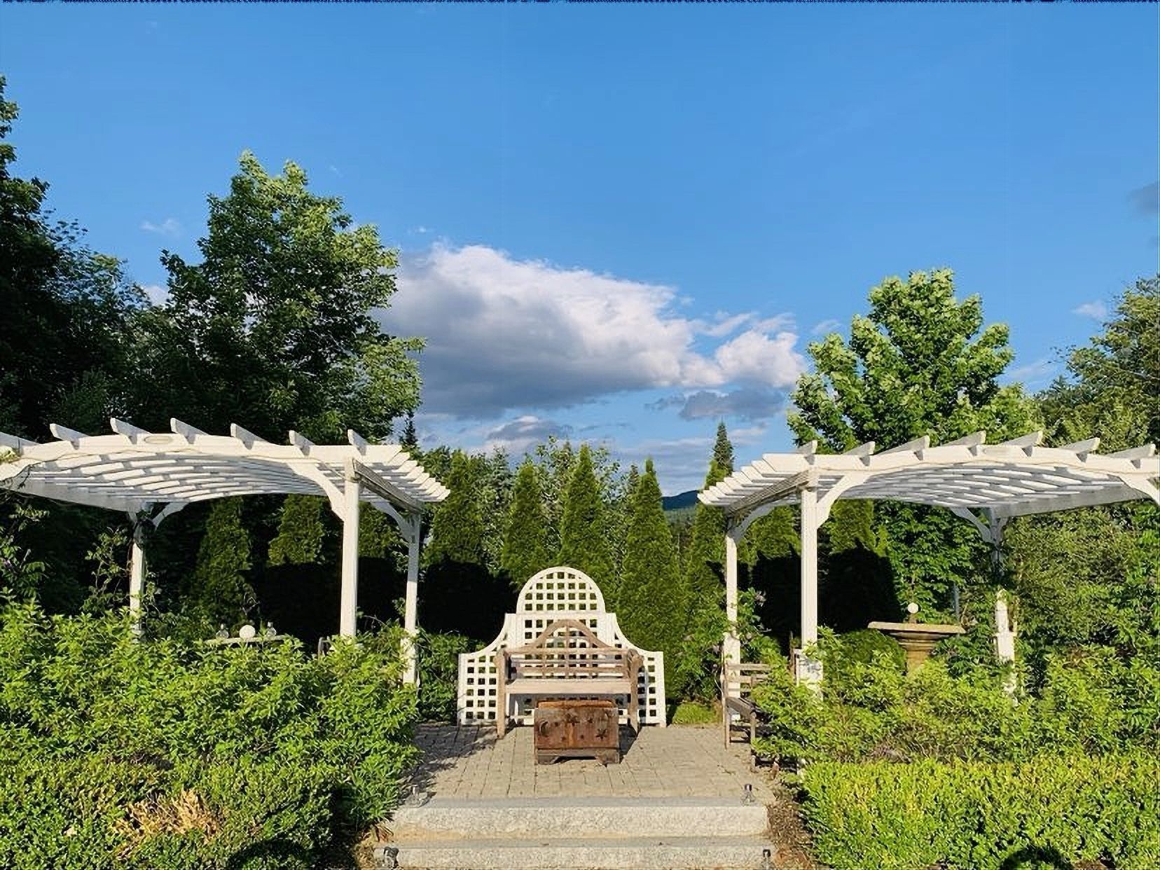 White pergola and bench in a garden, blue sky, trees, and greenery.