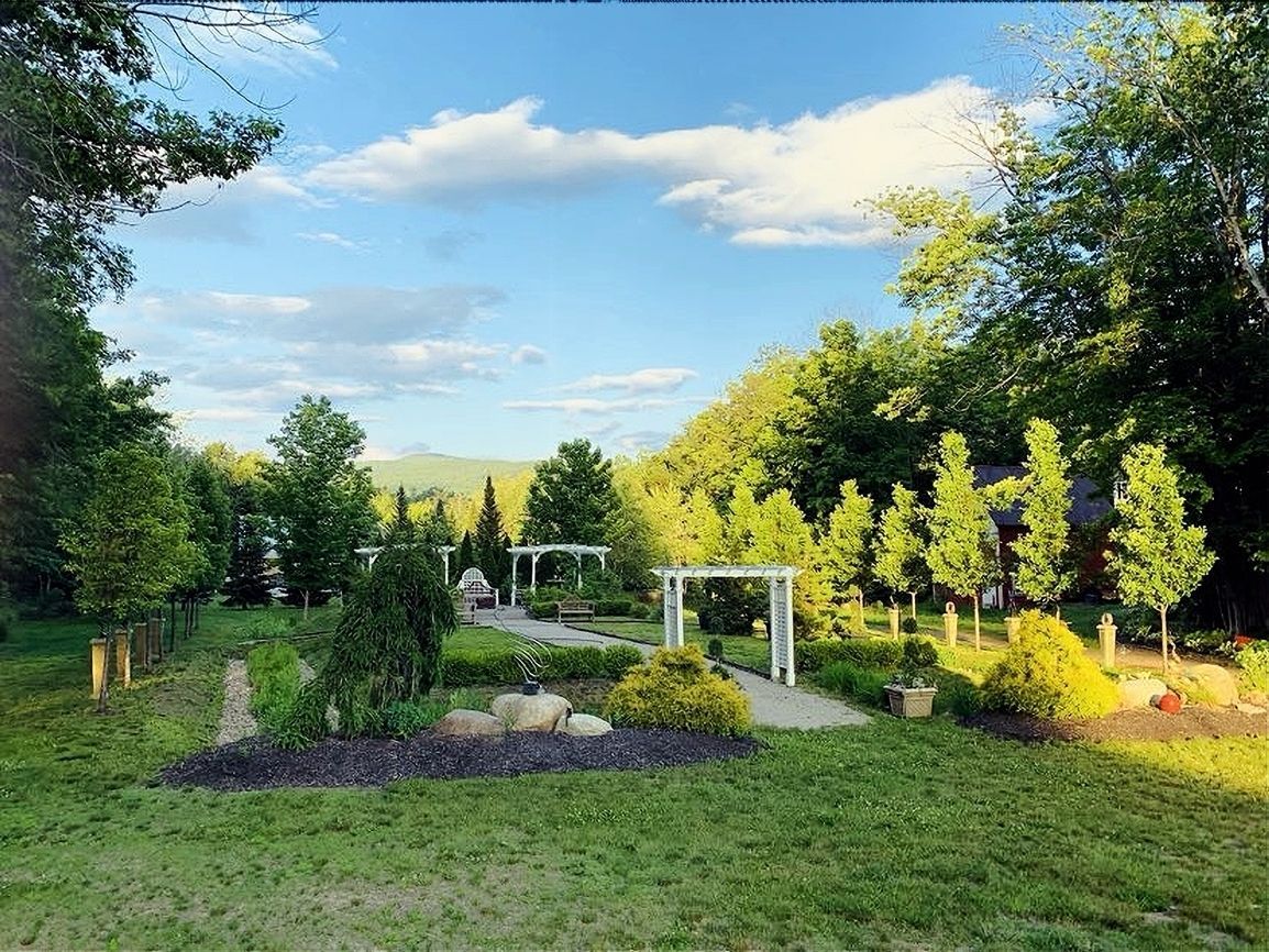 Lush green garden with manicured trees, white arches, and a mountain backdrop under a partly cloudy sky.