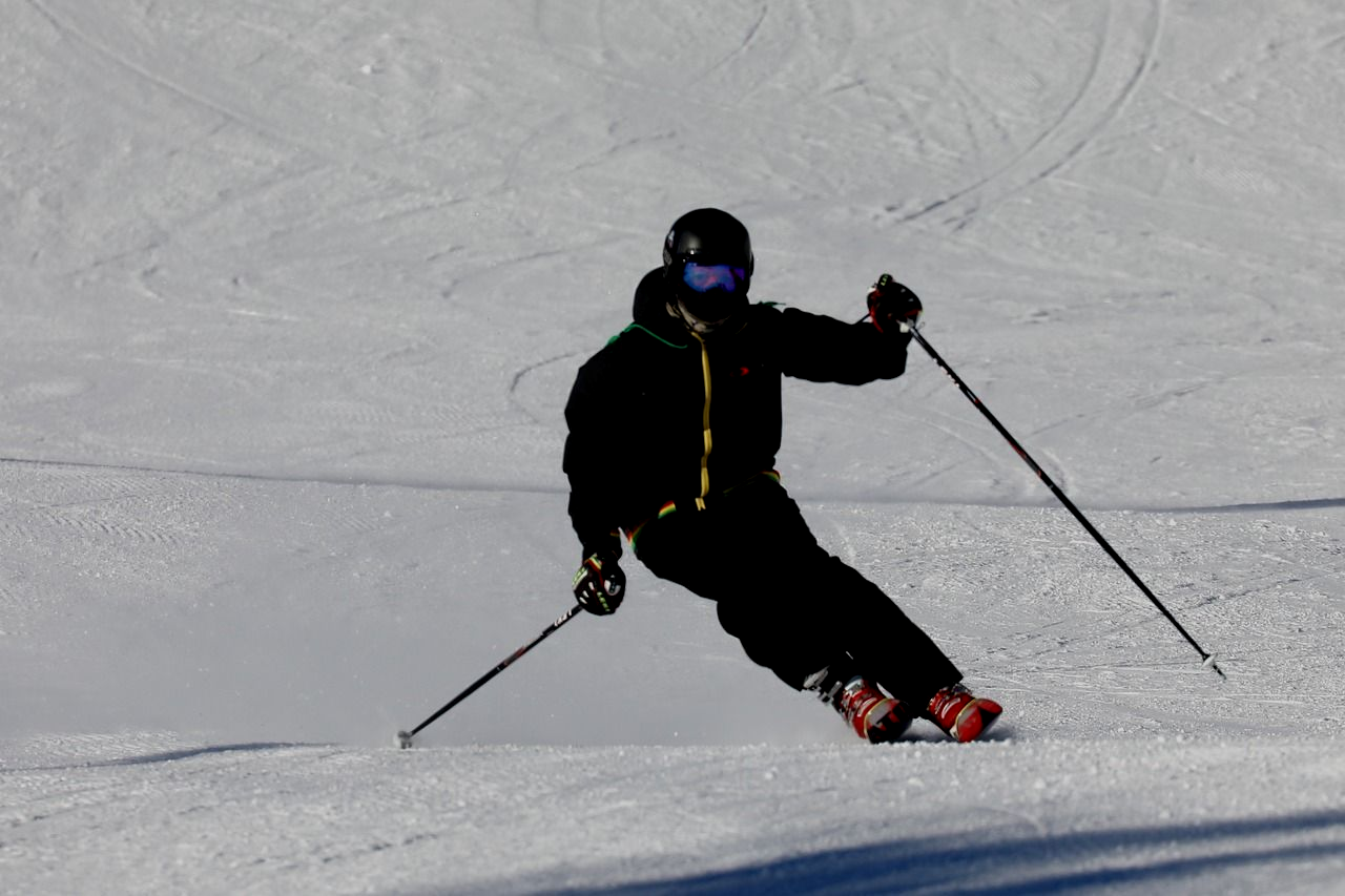 Skier in black gear carving down a snowy slope, poles extended, sunlit.