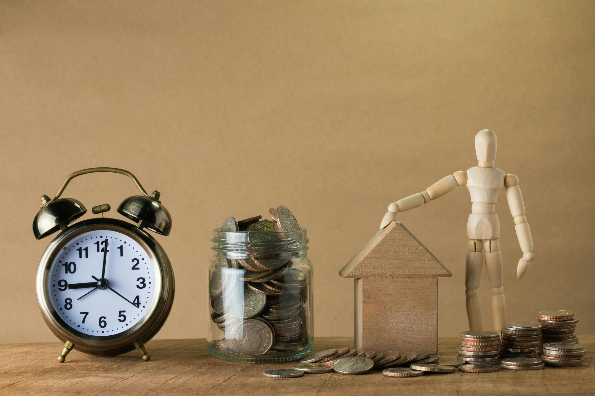 Alarm clock, jar of coins, small house model, and wooden mannequin on a beige background