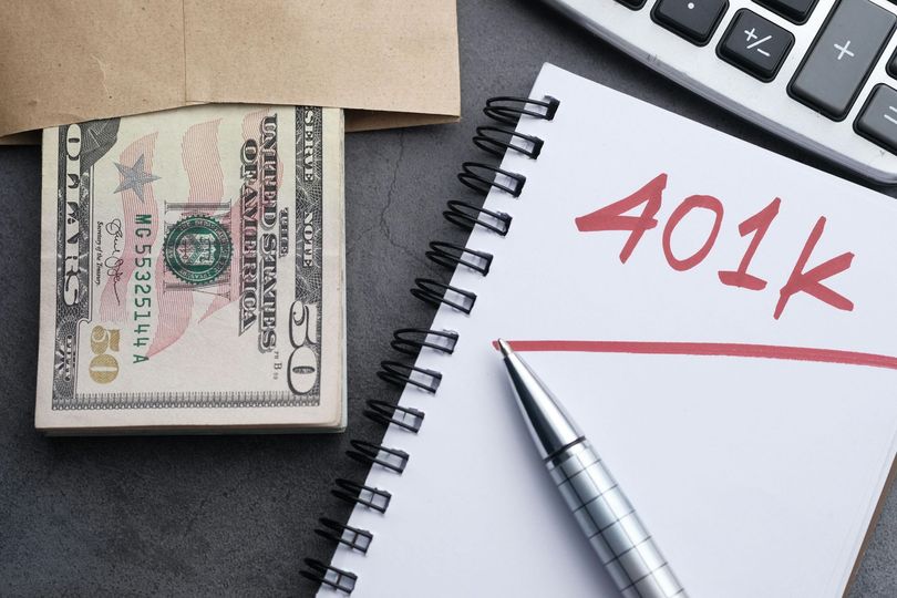$100 bill beside a notebook labeled “401k” with a pen on a desk