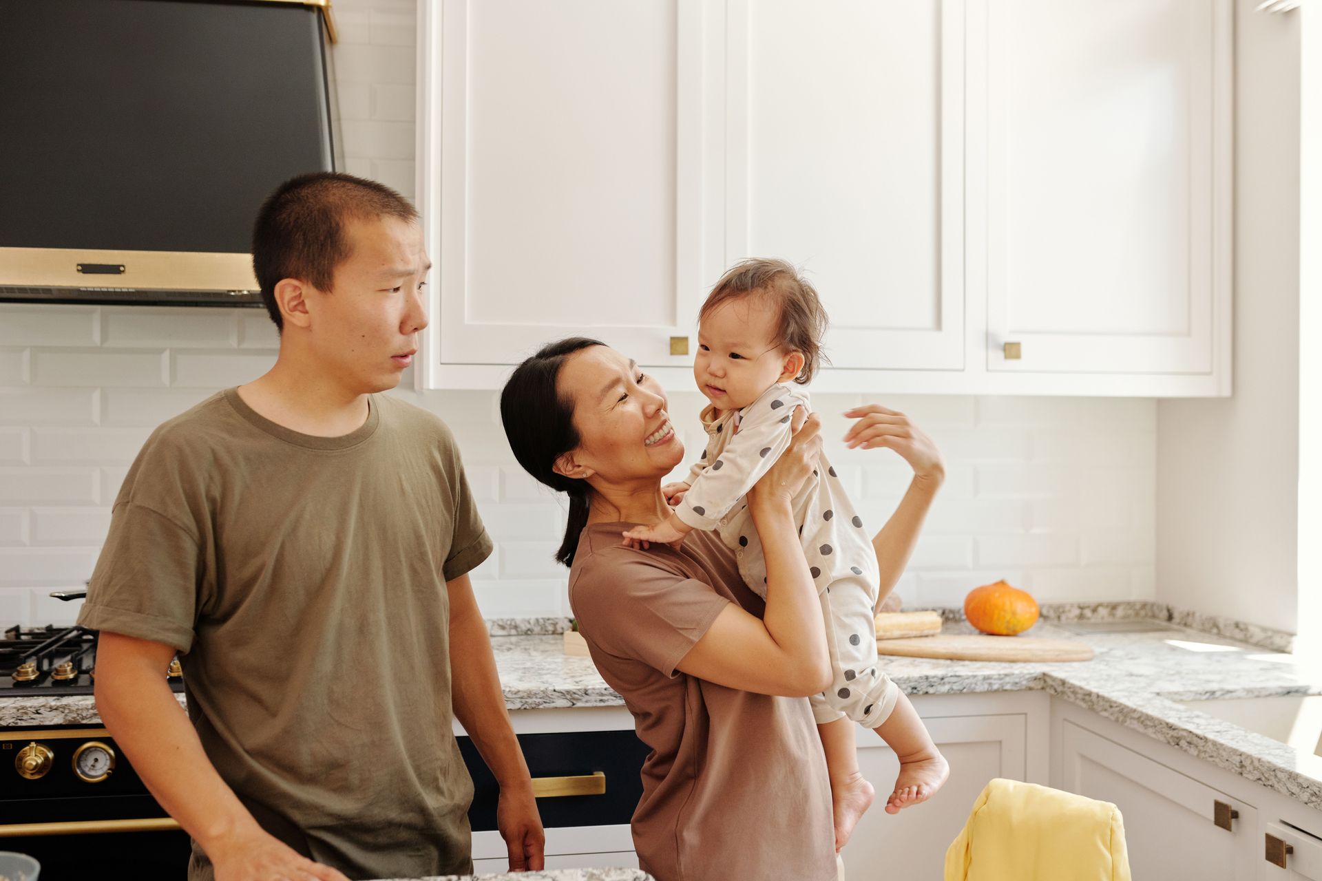 Three people in a bright kitchen, with one adult holding a child and another adult looking on.