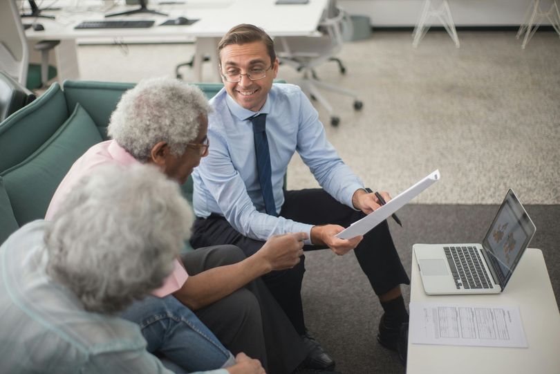 Three people in an office discuss a document beside an open laptop, with one man smiling and pointing.