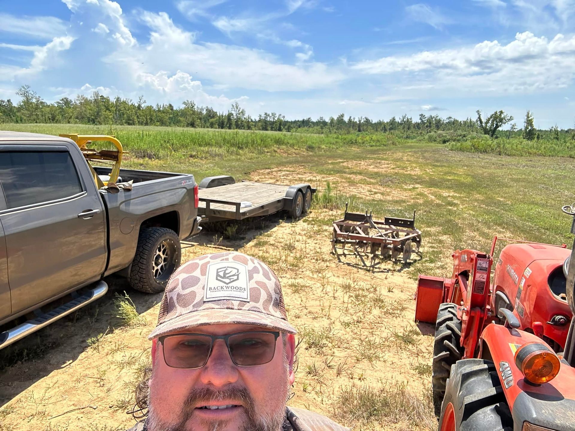 Man in camo hat taking a selfie outdoors with farm equipment. A truck, trailer, and tractor are visible in a sunny field.