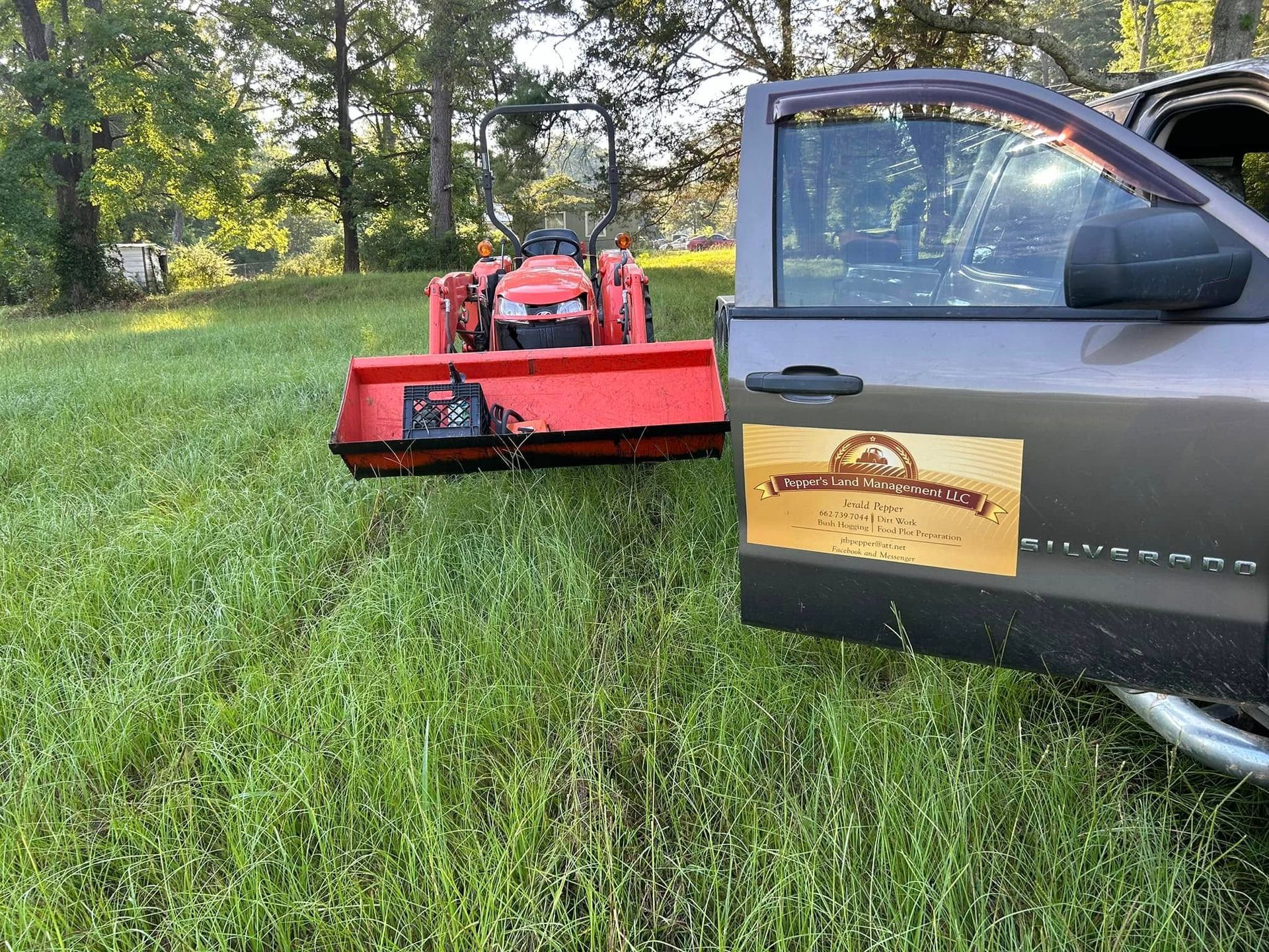A grey pickup truck parked in a grassy field has an open door. A tractor with a red mowing attachment is behind it.