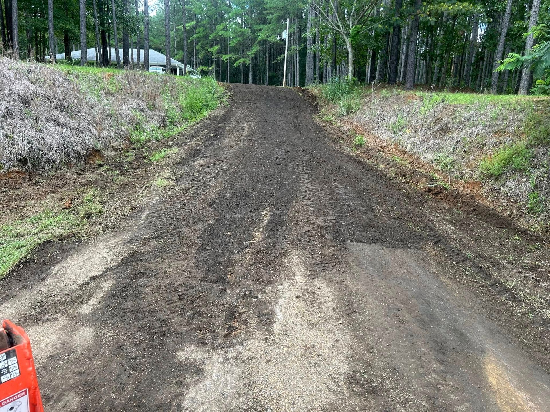 Dirt road with recent grading, flanked by grassy embankments and a treeline. A small section of a machine is visible on the bottom left.