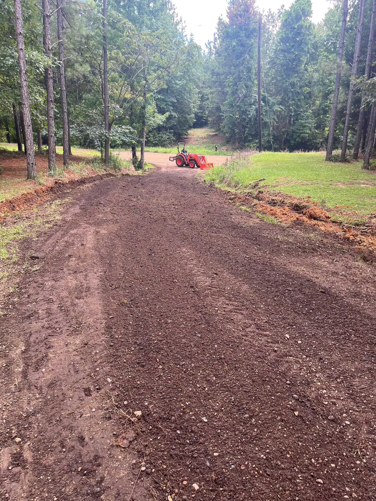 A garden bed, freshly tilled, in a grassy area bordered by trees. A tractor is visible in the background.