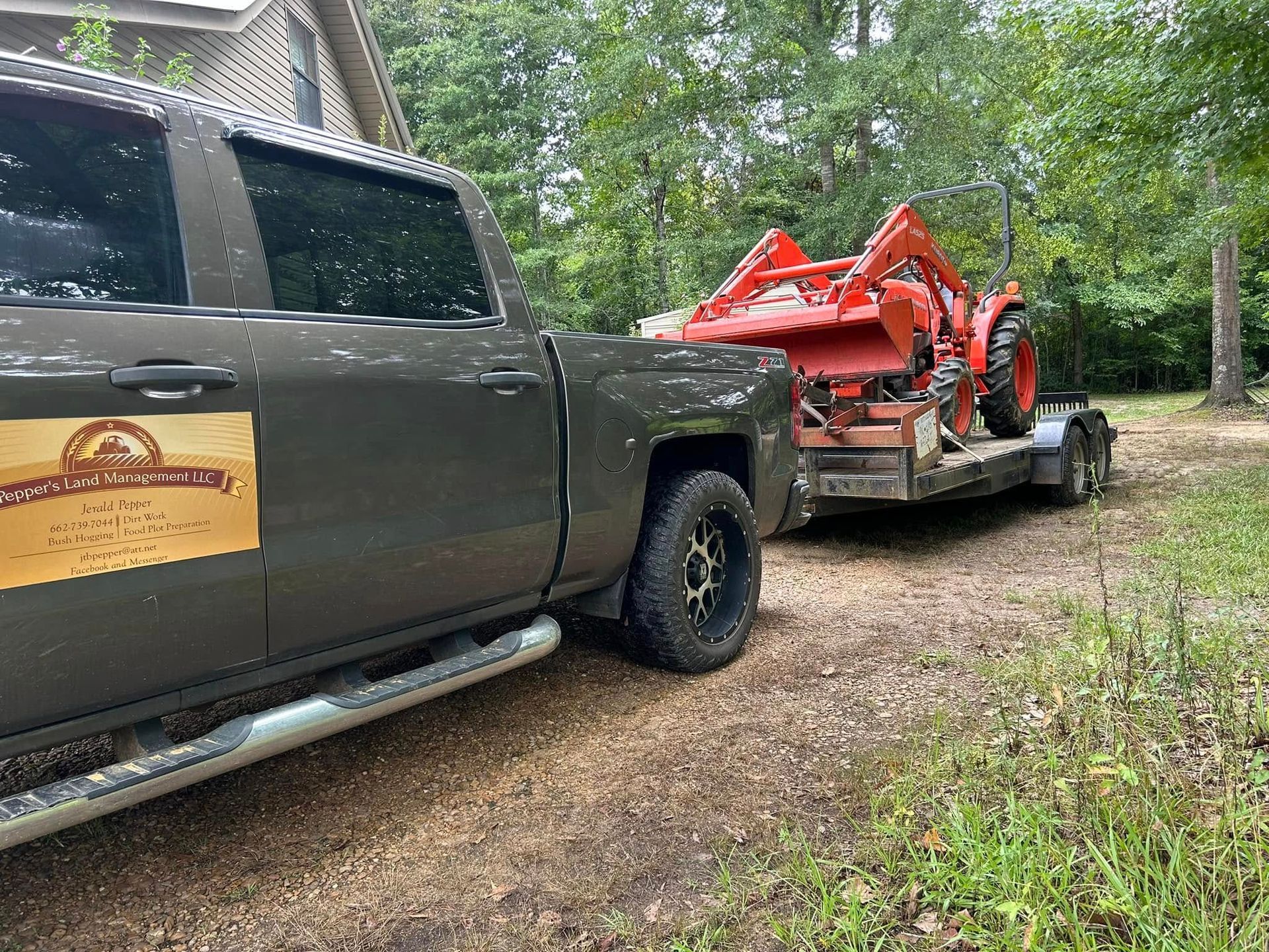 A gray pickup truck towing an orange tractor on a trailer on a dirt road.