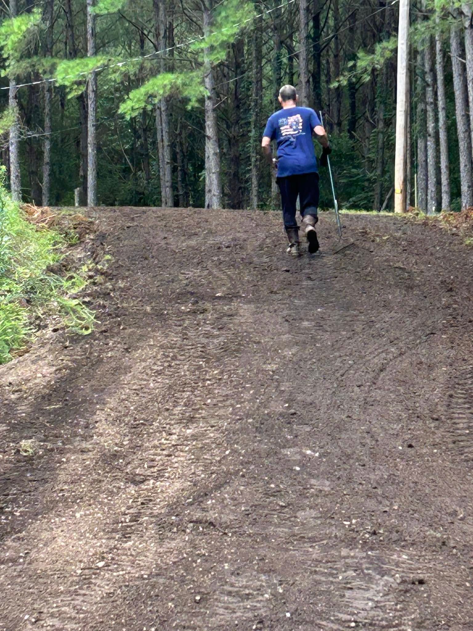 Person walking up a dirt hill in a forest setting, holding a pole.