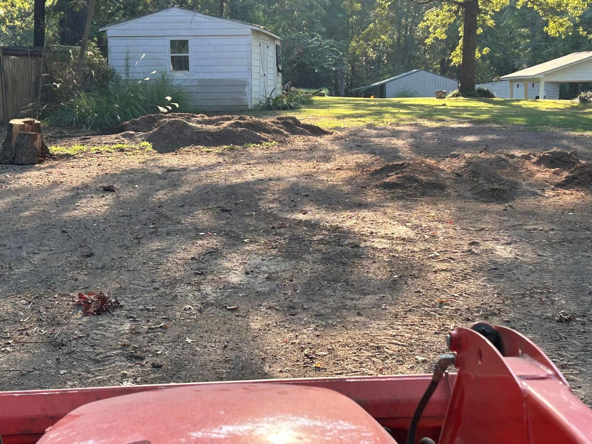 A red tractor in a gravel yard with dirt mounds, a white shed, and houses in the background.