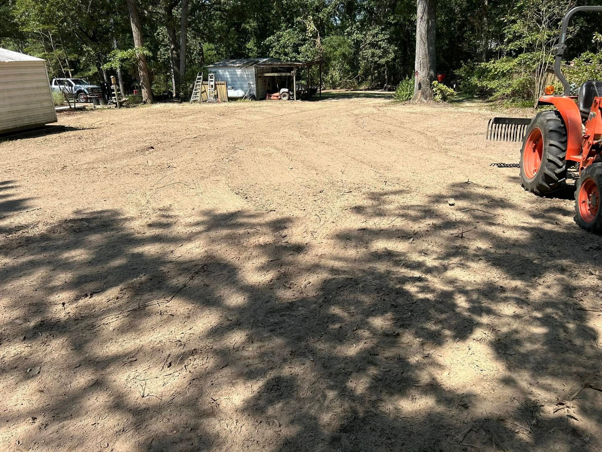 Tractor on a freshly graded gravel surface. Trees cast shadows, small structures and a building in the background.