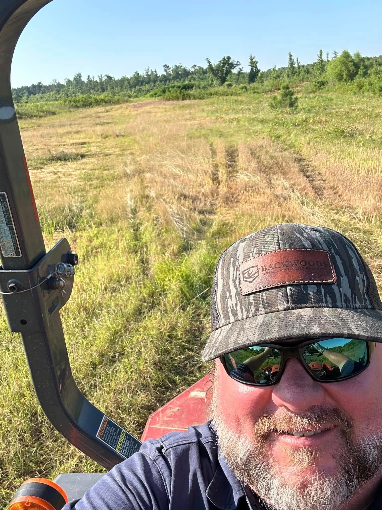 Man wearing sunglasses and a camo hat driving a tractor in a field.  He's smiling in the sun.