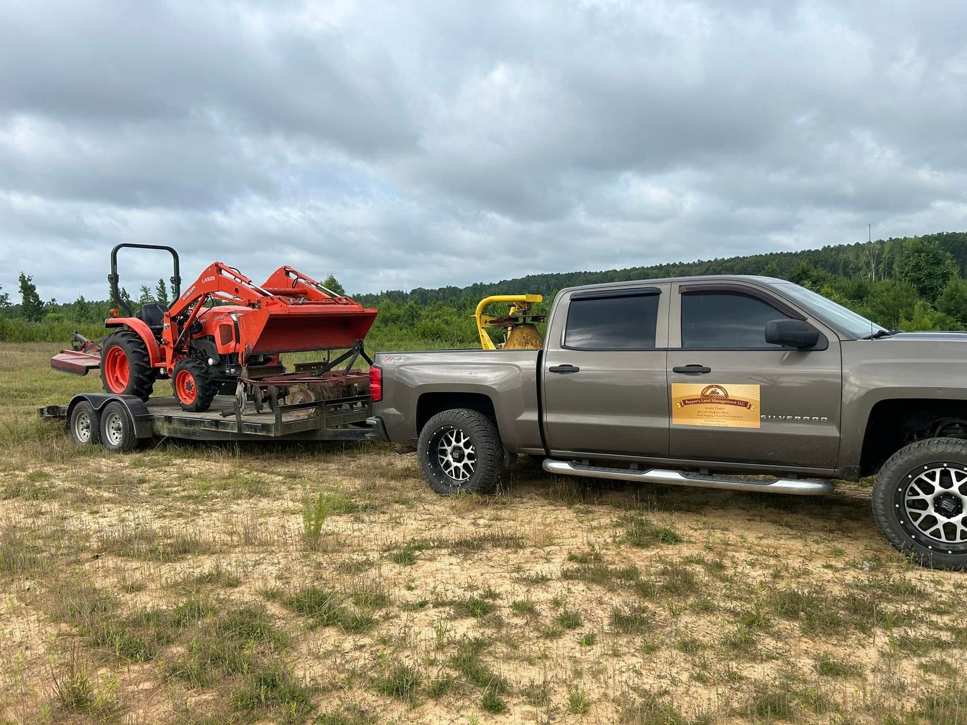 A brown pickup truck towing a trailer with an orange tractor on it; set in a grassy field with trees in the background.