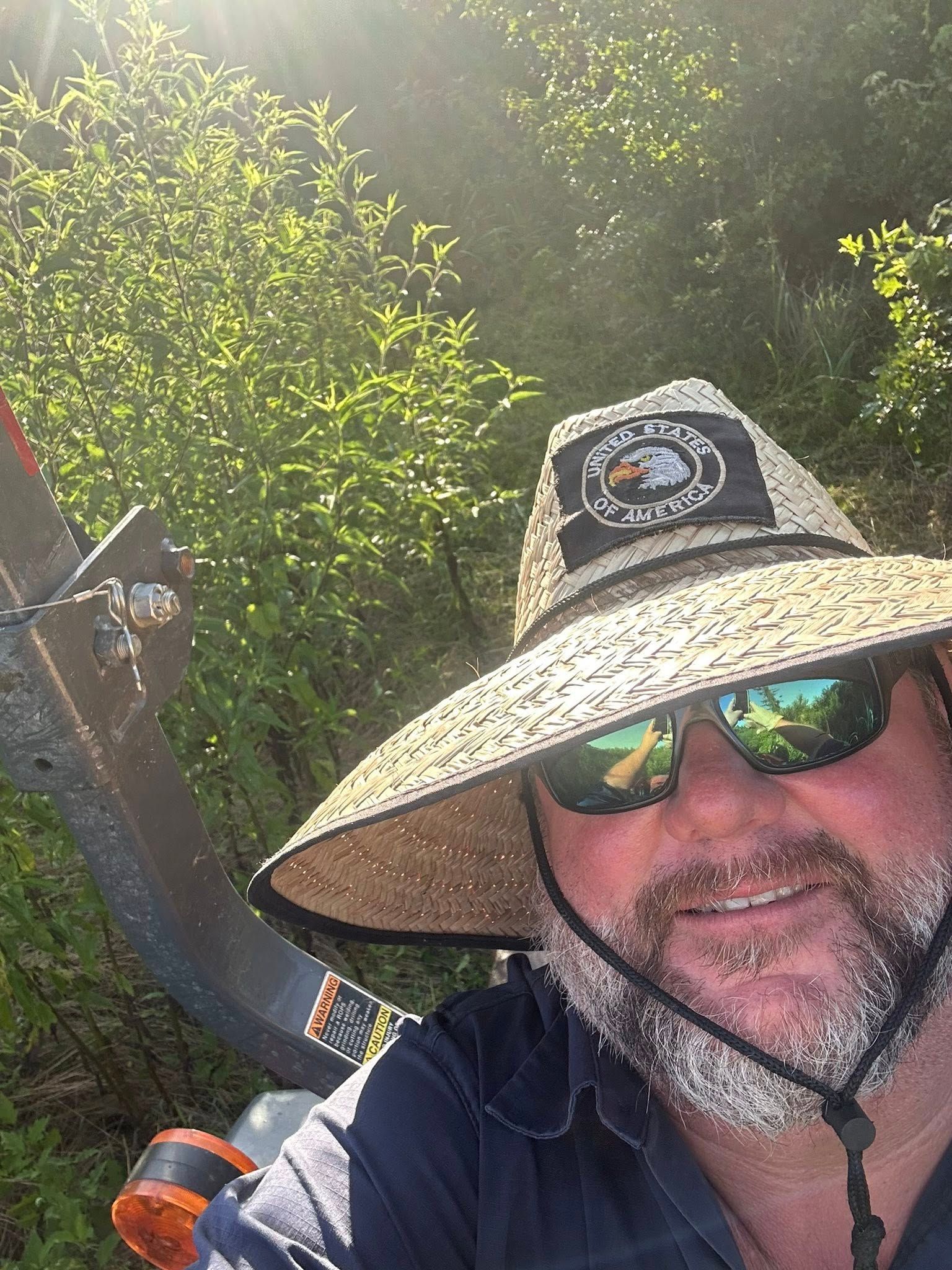 Man wearing a straw hat and sunglasses on a tractor in a sunny, green setting. He is smiling.