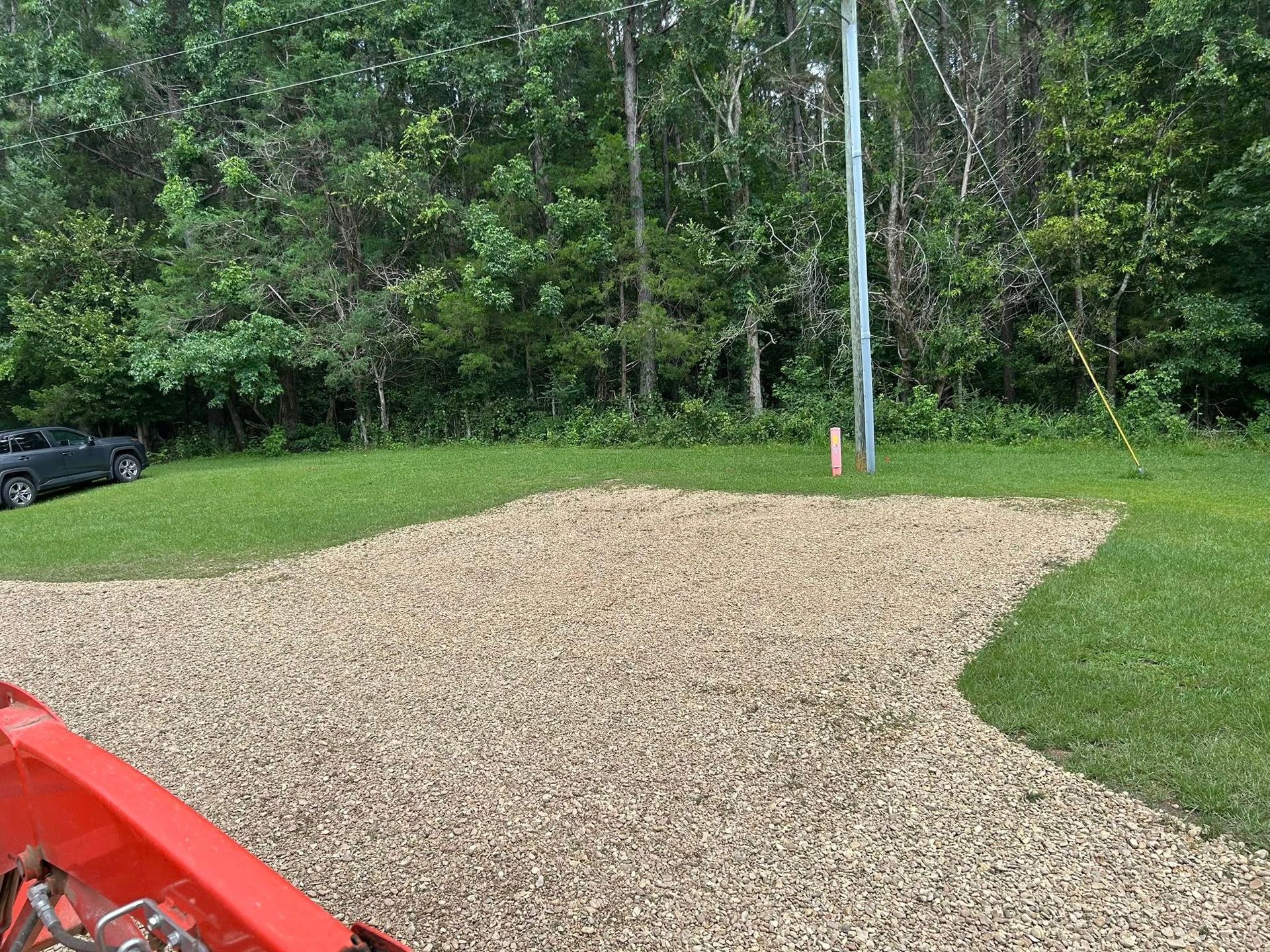 A gravel driveway bordered by green grass. Trees fill the background. A vehicle is parked to the left.