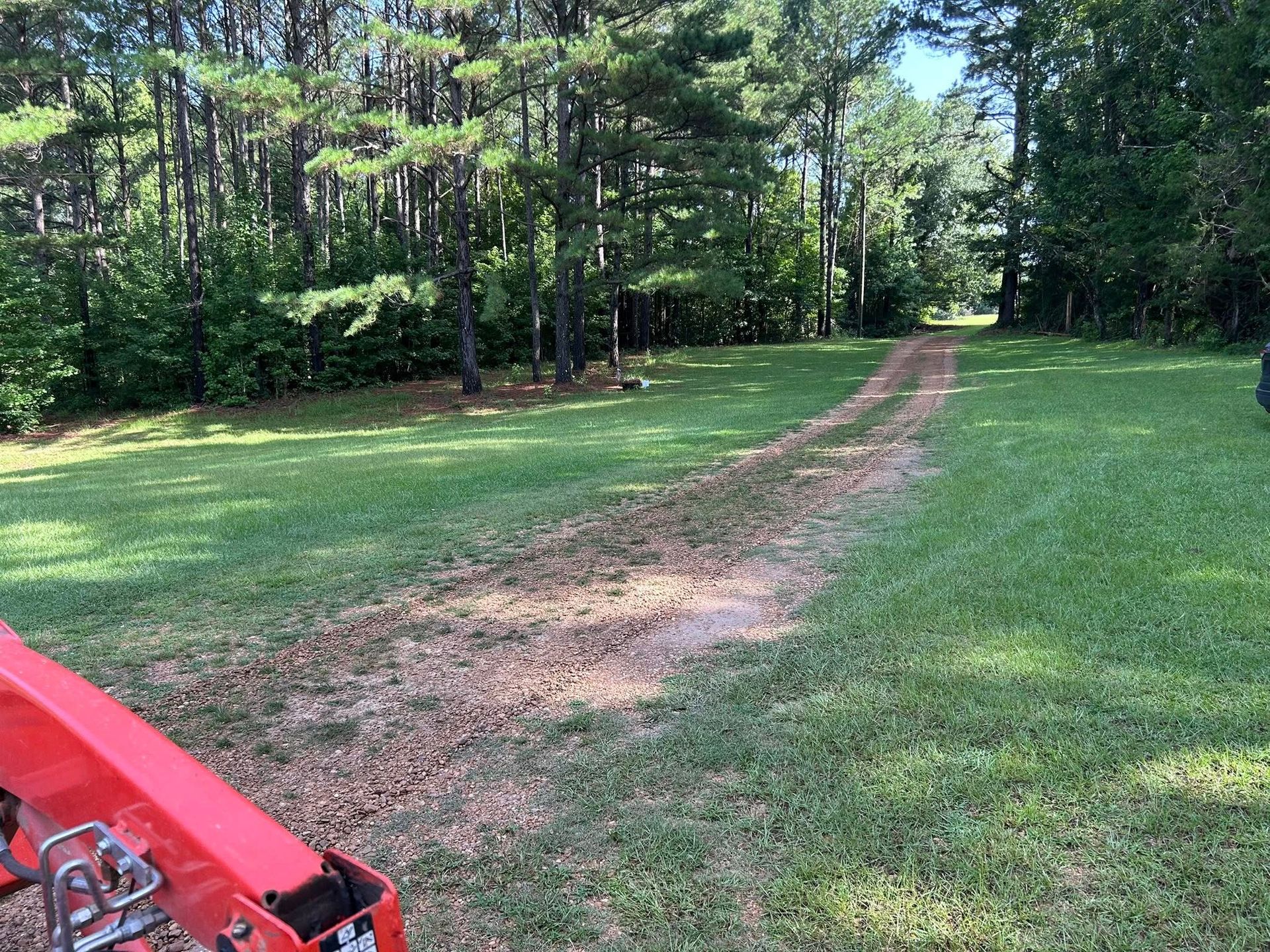 Dirt driveway through a grassy yard leading into a wooded area. A red tractor is in the foreground.