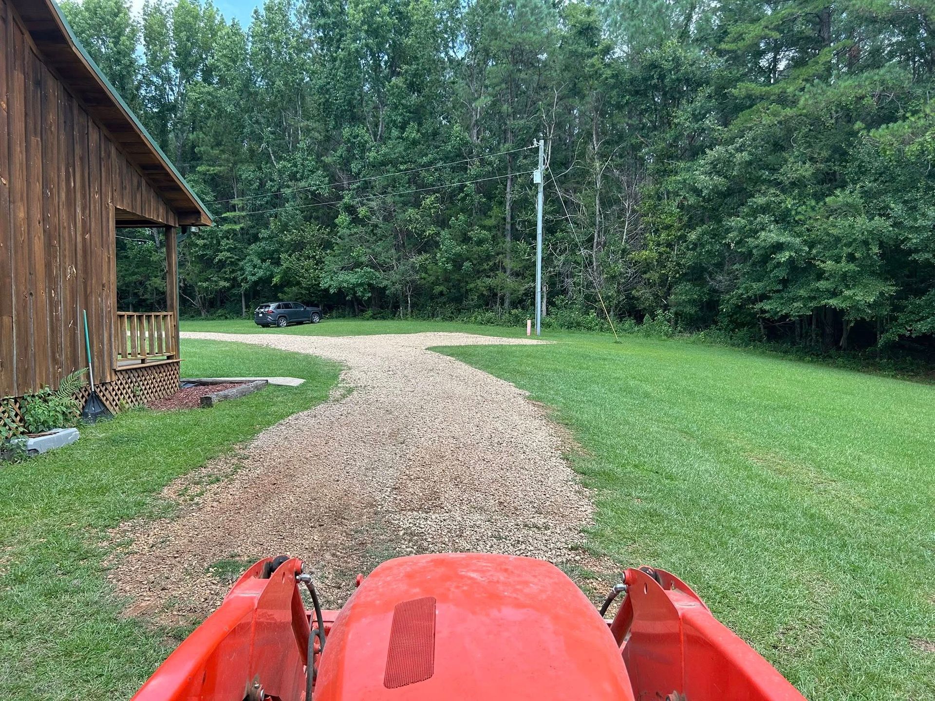 A tractor moving gravel on a driveway leading to a wooden house and trees on a grassy property.