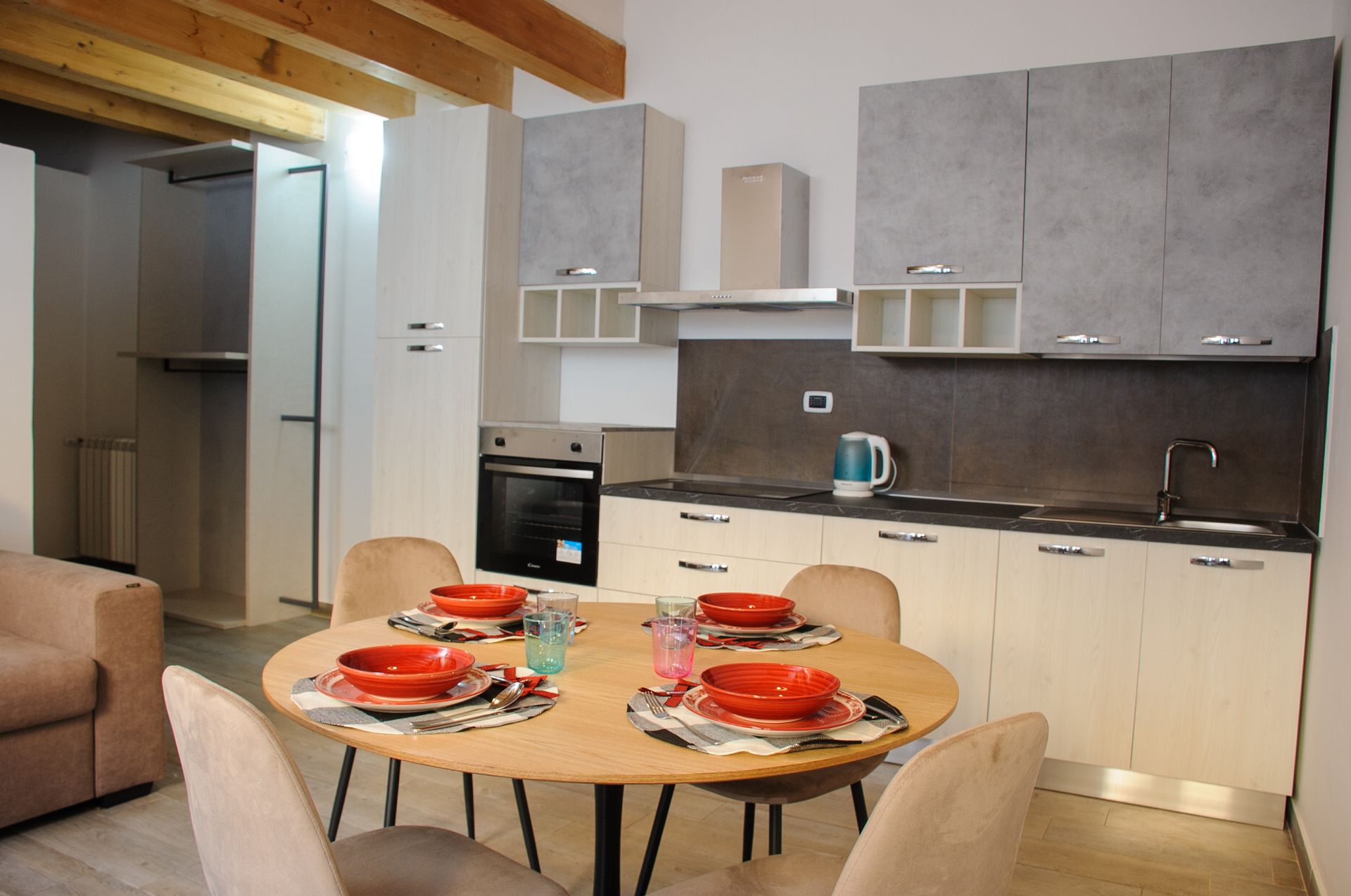 Cucina e zona pranzo moderne con tavolo rotondo per quattro persone, mobili grigi e travi in ​​legno a vista.