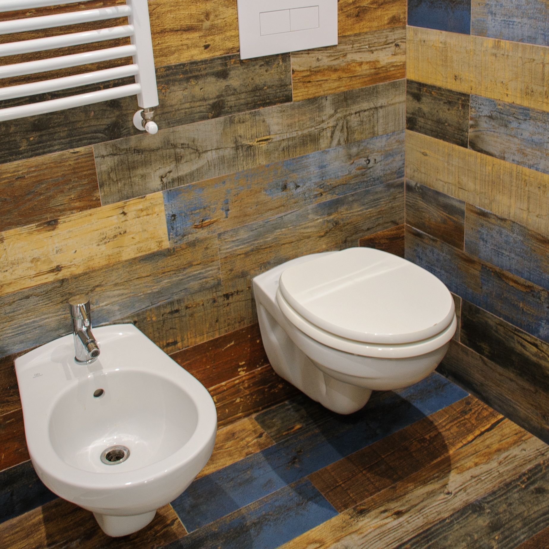Piccolo bagno con lavabo e WC a parete, caratterizzato da piastrelle colorate che riproducono l'effetto legno su pareti e pavimento.