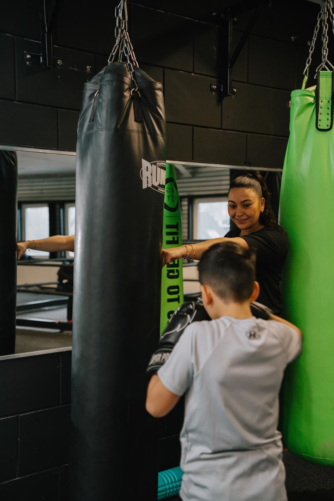 Een persoon die een kind begeleidt bij bokstraining, waarbij het kind op een zwarte, zware bokszak slaat in een sportschool.