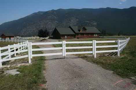 A white fence surrounds a dirt road leading to a house with mountains in the background.