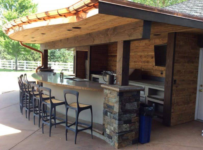 Outdoor bar with curved counter and seating. Features stone columns, wooden beams, and a copper rain gutter.