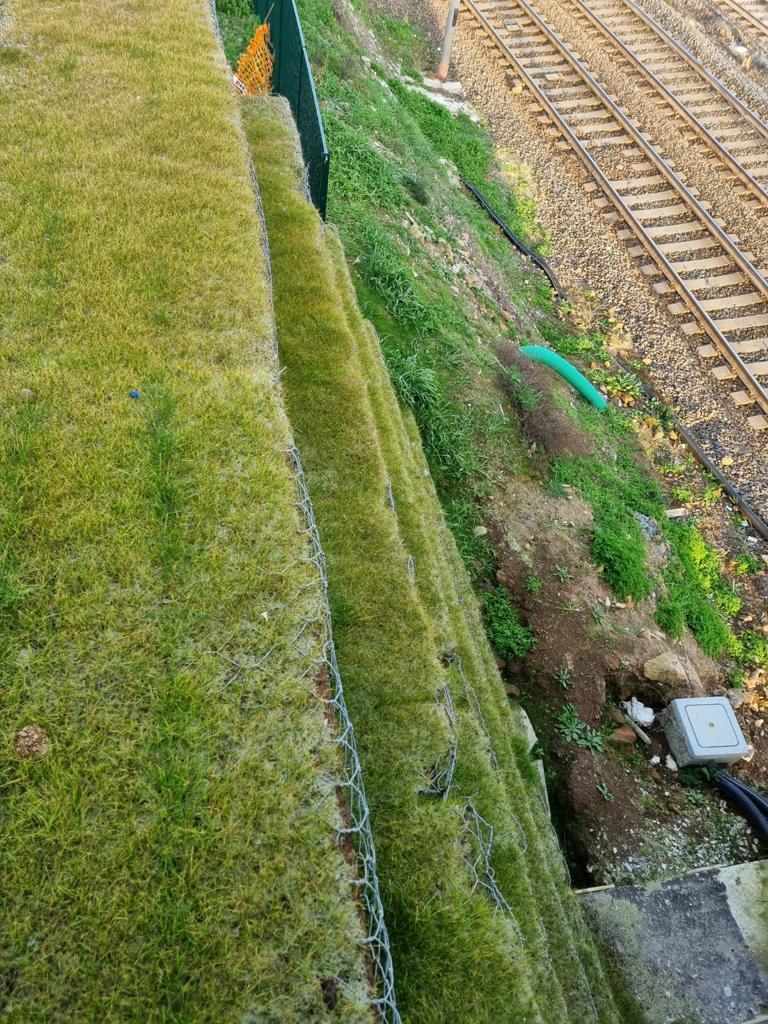 Vista dall'alto di un terrapieno verde terrazzato accanto ai binari della ferrovia, con una recinzione metallica e tubi di drenaggio.