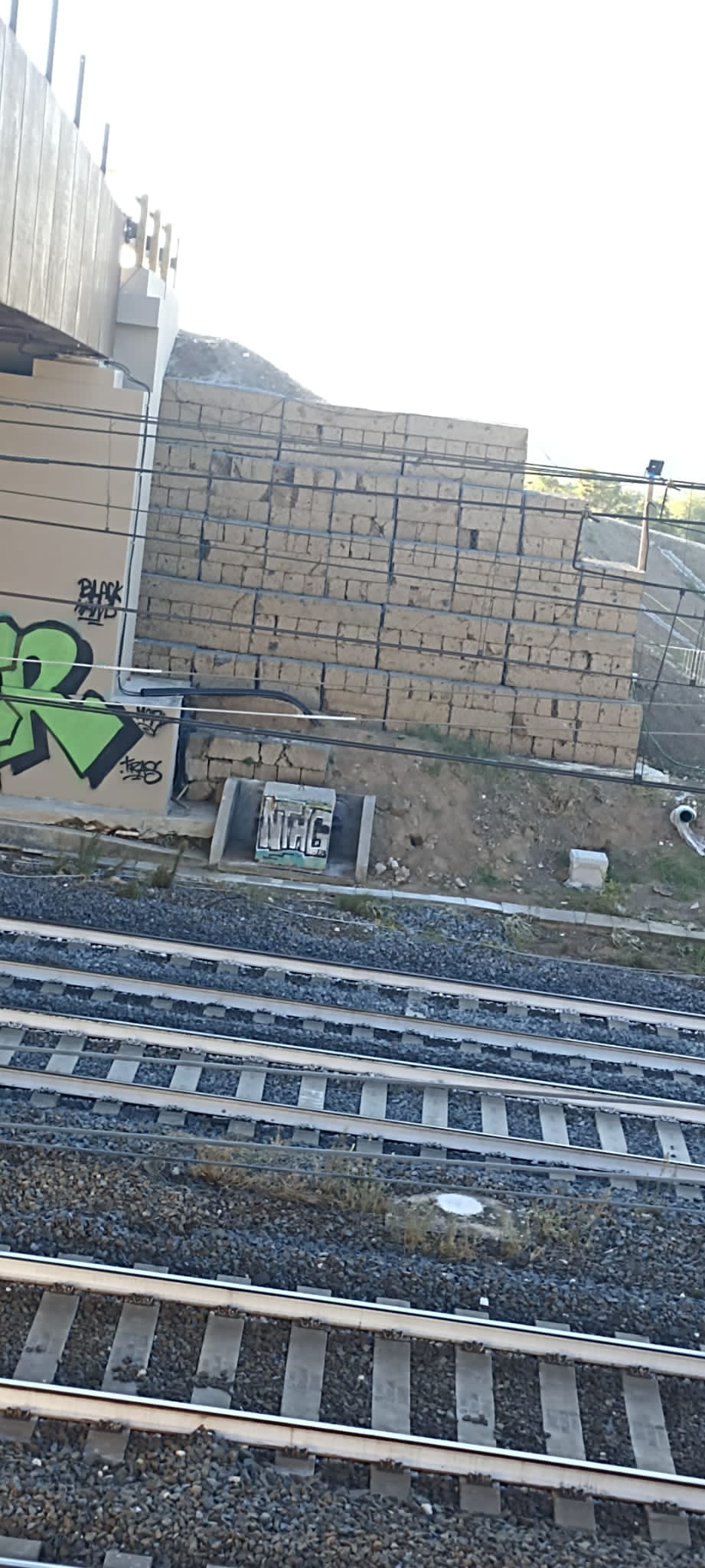 Una linea ferroviaria multi-binario accanto a un muro di cemento color marrone chiaro a più livelli, contrassegnato da graffiti verdi e da un piccolo cartello rettangolare.