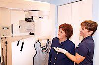 An older patient and dental assistant stand in front of an x-ray machine
