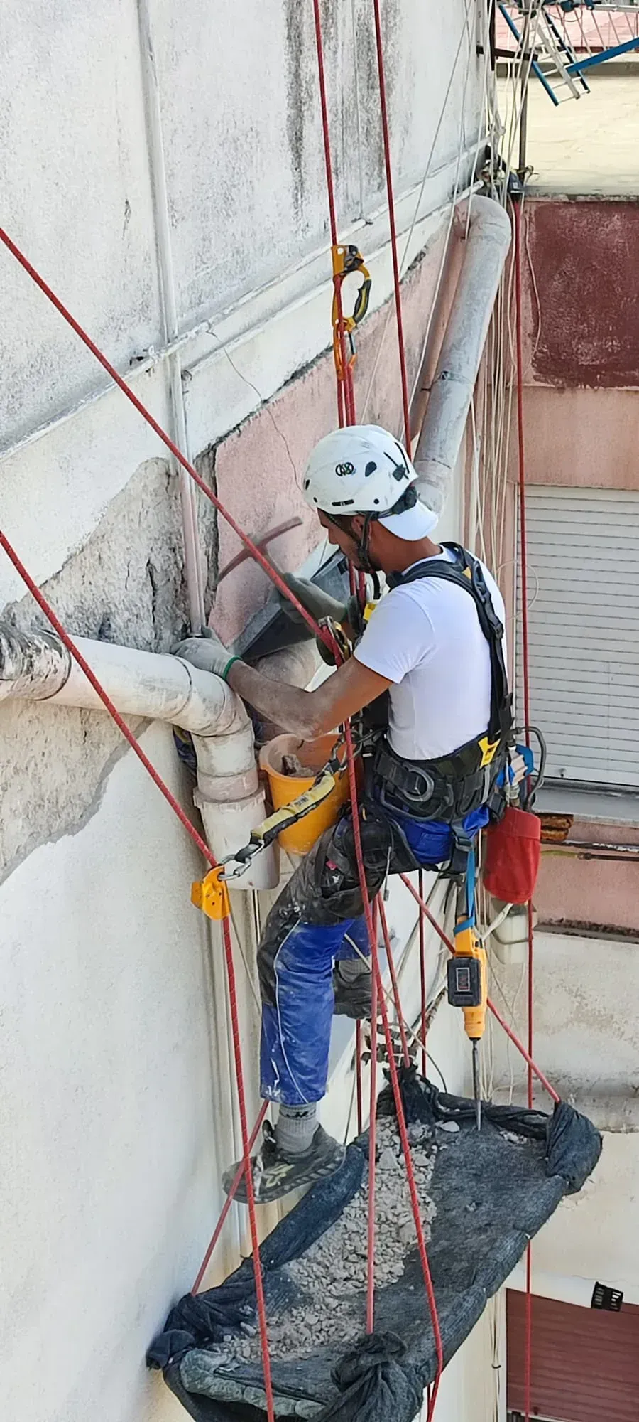 Uomo con equipaggiamento di sicurezza che lavora sul lato di un edificio, utilizzando degli attrezzi.