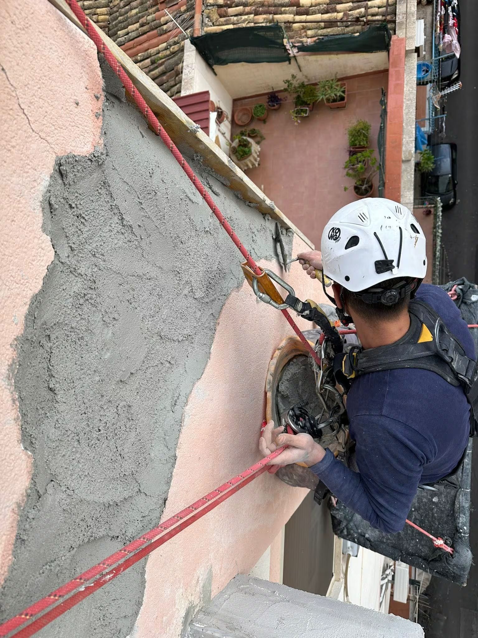 Operatore addetto all'accesso con funi su un edificio, che indossa casco e imbracatura, mentre ispeziona o lavora sul muro.