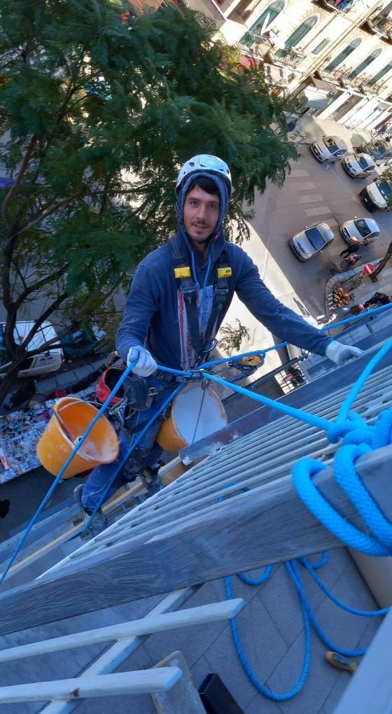 Tecnico di accesso su fune appeso al muro di un edificio. Indossa imbracatura, casco e tiene in mano gli attrezzi. Giornata di sole.