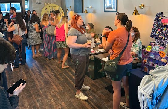 People line up at a table. A woman with red hair talks, a masked person sits at the table, a shop interior.